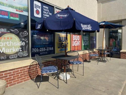 Outdoor seating area with tables and umbrellas in front of a market, featuring beer advertisements on the windows.