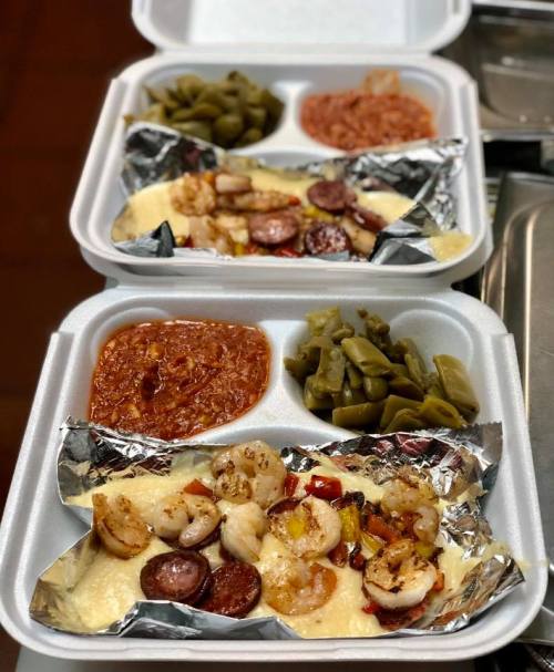 Two takeout containers with shrimp, sausage, and vegetables over cheese, accompanied by green beans and red sauce.