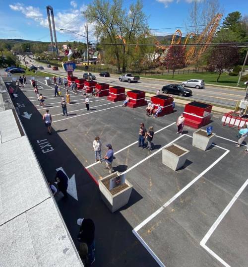 Aerial view of a parking lot with people, red seating areas, and roller coasters in the background on a sunny day.