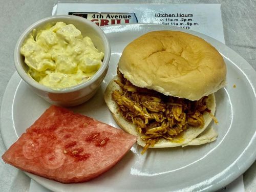 A plate with a pulled pork sandwich on a bun, a side of potato salad, and a slice of watermelon.