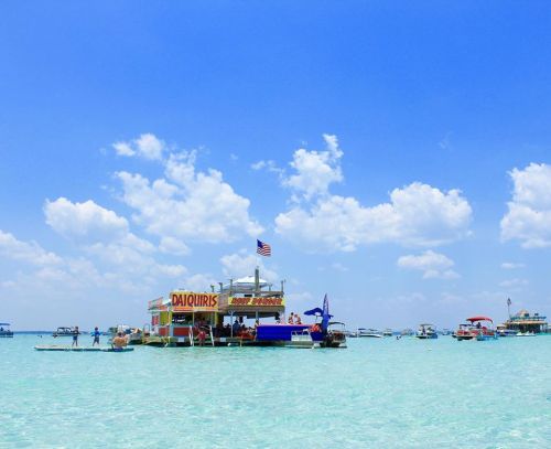 A colorful floating bar with an American flag, surrounded by clear blue water and boats under a bright sky.
