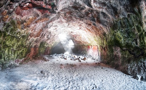 A cavern with colorful rock walls and a sandy floor, illuminated by a beam of light from an opening above.