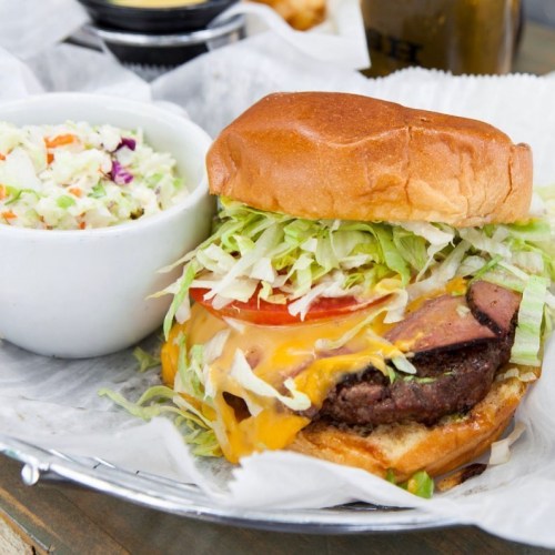 A juicy cheeseburger with lettuce, tomato, and a side of coleslaw in a white bowl on a rustic table.