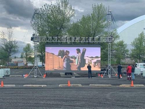 Outdoor screen showing a scene from a film, with people watching in a parking lot surrounded by trees.