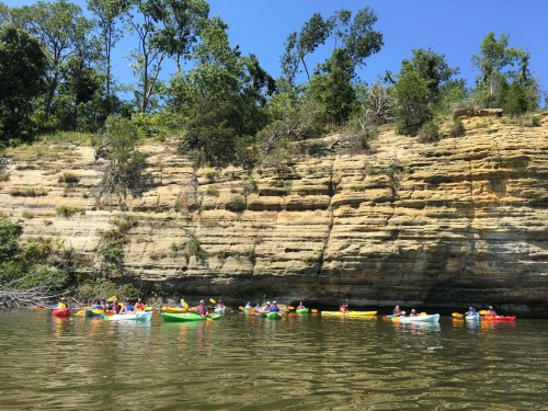 A group of kayakers paddles along a river with steep, rocky cliffs and lush greenery in the background under a clear blue sky.