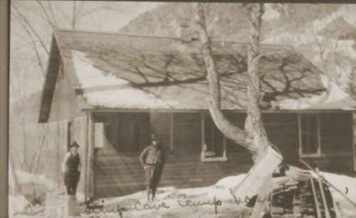 Two men stand outside a rustic cabin in a snowy landscape, with trees casting shadows on the building.