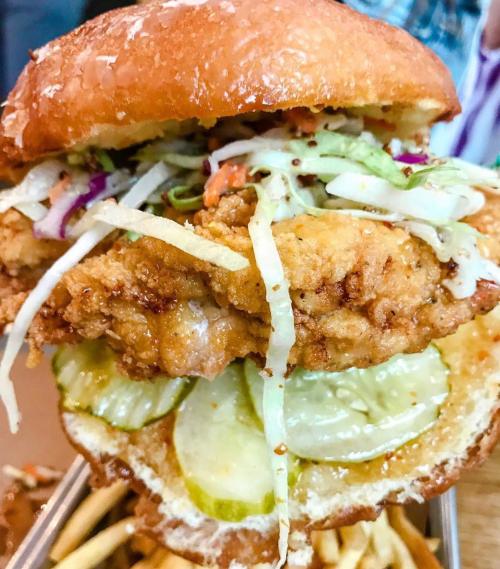 A towering fried chicken sandwich with pickles, slaw, and sauce, served with a side of fries.