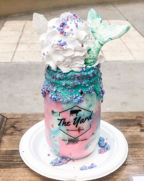 A colorful milkshake in a jar topped with whipped cream, sprinkles, and a mermaid tail decoration.