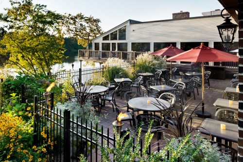 Outdoor dining area with tables, umbrellas, and lush greenery by a lake, featuring a cozy, inviting atmosphere.