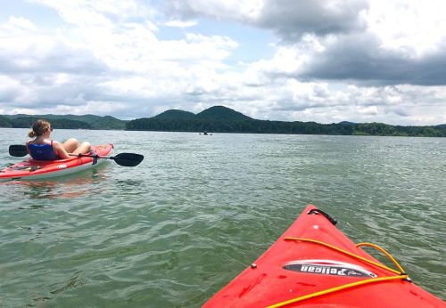 Two kayakers on a calm lake, surrounded by green hills and cloudy skies. One person relaxes in their kayak.