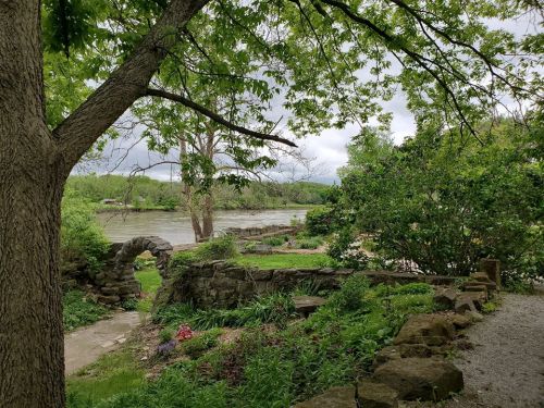 A serene riverside scene with lush greenery, stone pathways, and a cloudy sky in the background.