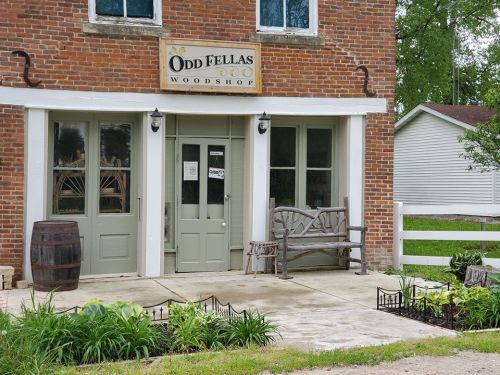 Exterior of Odd Fellas Woodshop, featuring a brick facade, green doors, a wooden bench, and a barrel by the entrance.