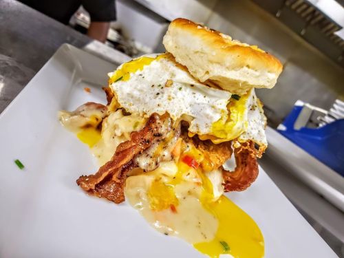 A towering breakfast dish with a biscuit, fried egg, bacon, and creamy sauce, served on a white plate.