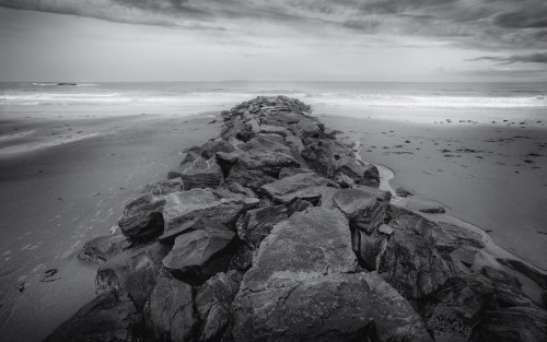A rocky jetty extends into a calm ocean under a cloudy sky, with gentle waves lapping at the shore.