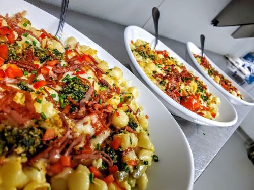 Three bowls of pasta topped with bacon, tomatoes, chives, and pesto, arranged on a stainless steel surface.