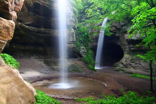 A serene landscape featuring two waterfalls cascading into a rocky area surrounded by lush green foliage.