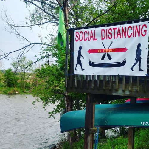 A sign promoting social distancing near a river, featuring silhouettes of people and a kayak, with "6 FEET" displayed.