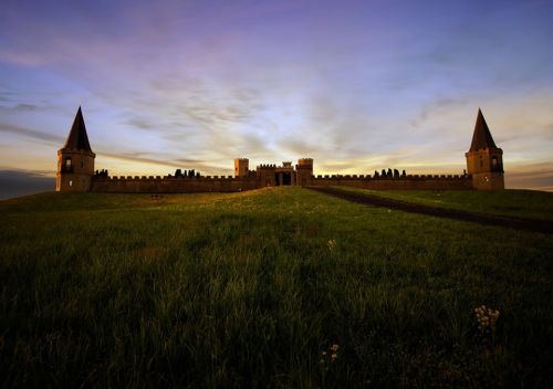A majestic castle with two tall towers, set against a colorful sunset sky, surrounded by green fields.