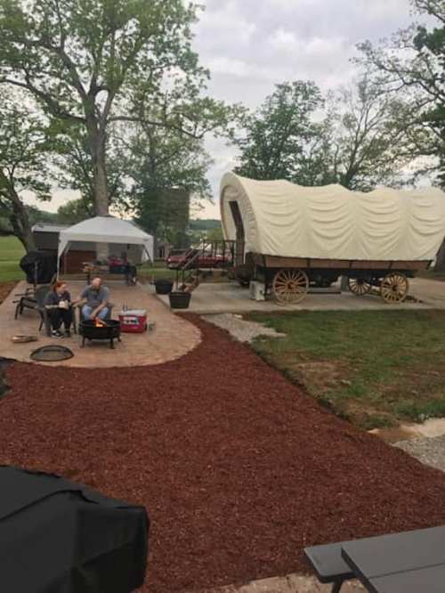A campsite with a covered wagon, fire pit, and seating area surrounded by trees and gravel paths.