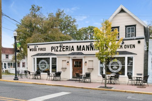 Exterior of Pizzeria Manna, featuring a wood-fired oven, outdoor seating, and a charming white building.