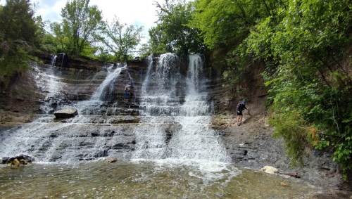 Two people explore a cascading waterfall surrounded by lush greenery and rocky terrain.