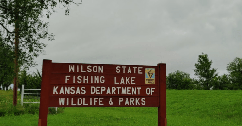 Sign for Wilson State Fishing Lake, Kansas Department of Wildlife & Parks, with green grass and cloudy sky in the background.