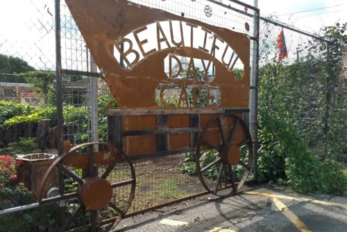 Rusty metal sign reading "Beautiful Day Café" with a garden backdrop and a chain-link fence.