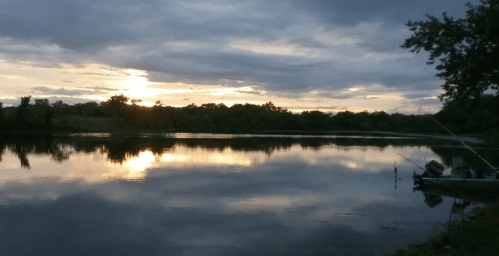 A serene sunset over a calm lake, with trees in the background and a fishing boat at the water's edge.
