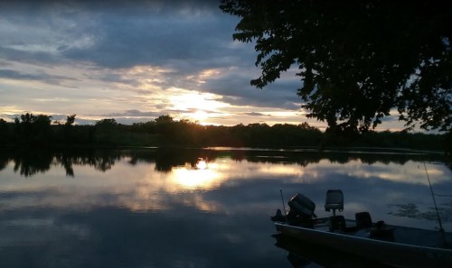 A serene sunset over a calm lake, with a boat docked at the shore and trees lining the water's edge.