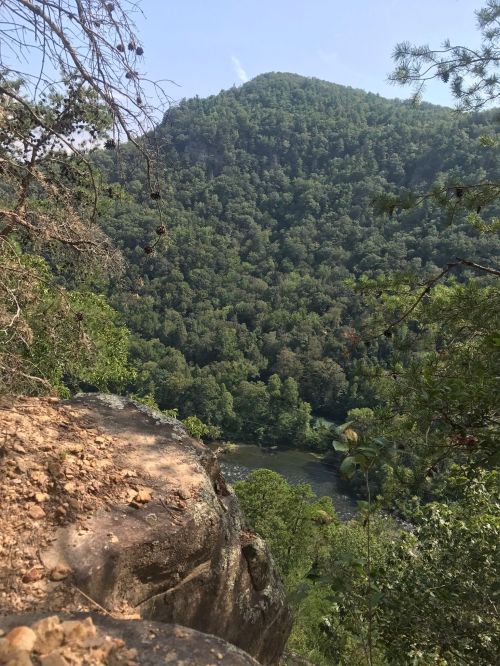 A rocky ledge overlooks a lush green valley and river, framed by trees and a distant mountain under a clear sky.