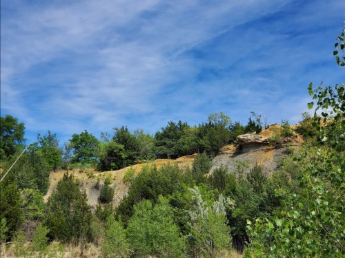 A scenic view of a rocky cliff surrounded by lush greenery and trees under a bright blue sky with wispy clouds.