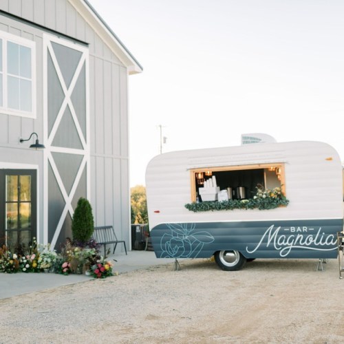 A vintage-style trailer bar named "Bar Magnolia" parked beside a barn, adorned with flowers and greenery.