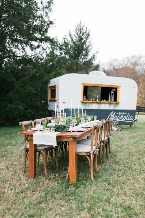 A rustic outdoor dining setup with a wooden table, chairs, and a vintage trailer in the background.