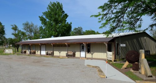 A single-story building with a metal roof, surrounded by trees and gravel, featuring multiple entrances and outdoor seating.