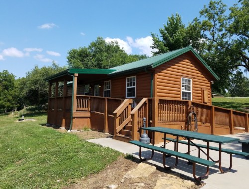 A brown log cabin with a green roof, surrounded by grass and trees, featuring a porch and picnic table.