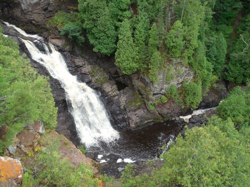 A cascading waterfall flows through rocky terrain, surrounded by lush green trees and vegetation.