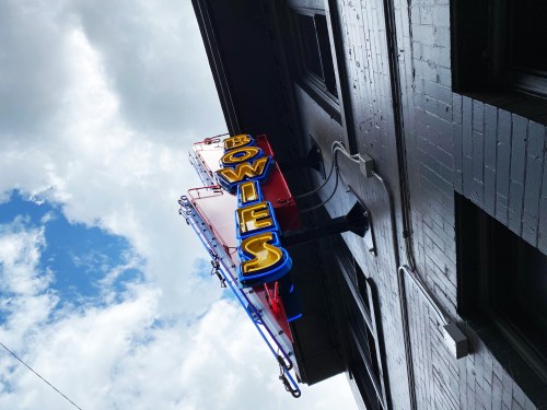 A colorful neon sign reading "Bowies" on a building, set against a cloudy blue sky.
