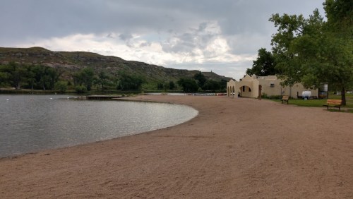 A sandy beach by a calm lake, surrounded by green trees and hills under a cloudy sky.