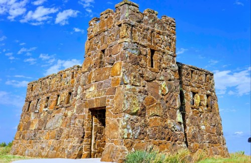 A stone fortress with a square tower against a blue sky, surrounded by grass and a clear landscape.
