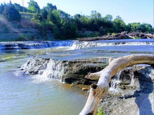 A serene waterfall cascades over rocky ledges, surrounded by lush greenery and a fallen log in the foreground.