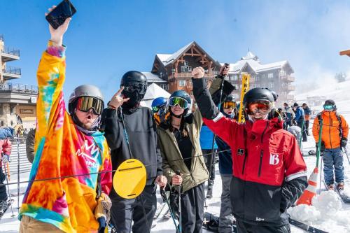 A group of five snowboarders in colorful gear, smiling and celebrating on a sunny ski slope.