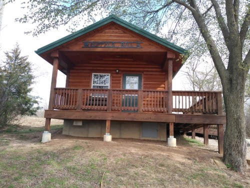 A wooden cabin with a green roof, front porch, and surrounding trees, set on a grassy area.