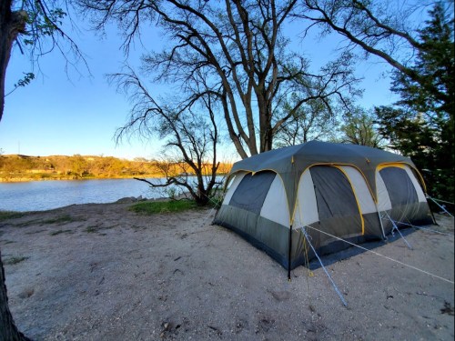 A tent set up by a calm lake, surrounded by trees and a clear blue sky.