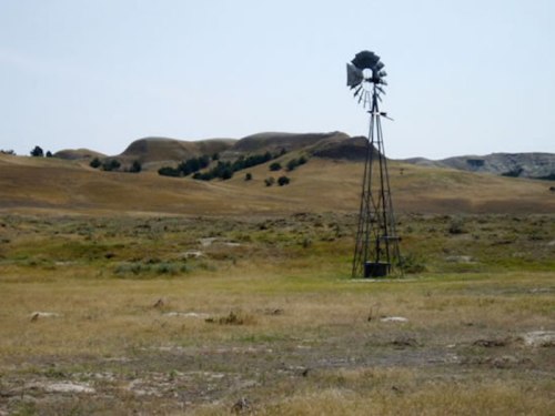 A tall windmill stands in a vast, dry landscape with rolling hills in the background under a clear sky.