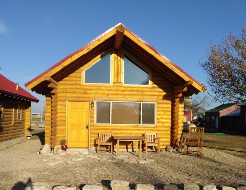 A cozy log cabin with large windows, a wooden door, and a stone patio, surrounded by a grassy area and blue sky.