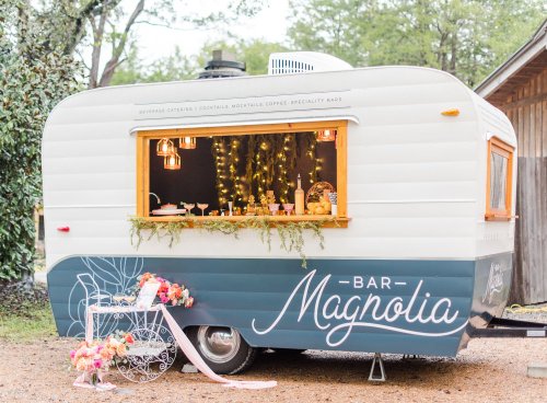 A vintage trailer bar named "Bar Magnolia" with decorative lights and floral arrangements, set in a natural outdoor setting.