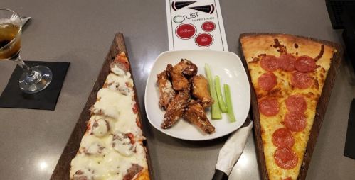 A table with two slices of pizza, chicken wings, celery sticks, and a drink, with a menu in the background.