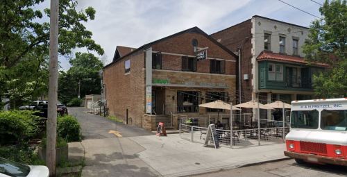 A brick building with outdoor seating, surrounded by trees and parked cars, on a sunny day.