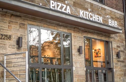 Exterior of a restaurant named "Pizza Kitchen Bar," featuring large windows and a stone facade.