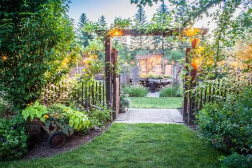 A lush garden pathway leads through a wooden archway adorned with lights, surrounded by vibrant greenery and flowers.